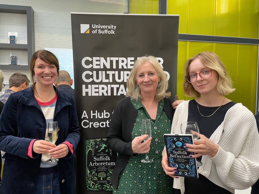 A photo of the editing team for the Suffolk Reflections volume at the launch event of the book standing by a banner for the school. From left to right is Lindsey Scott, Amanda Hodgkinson and Amber Spalding
