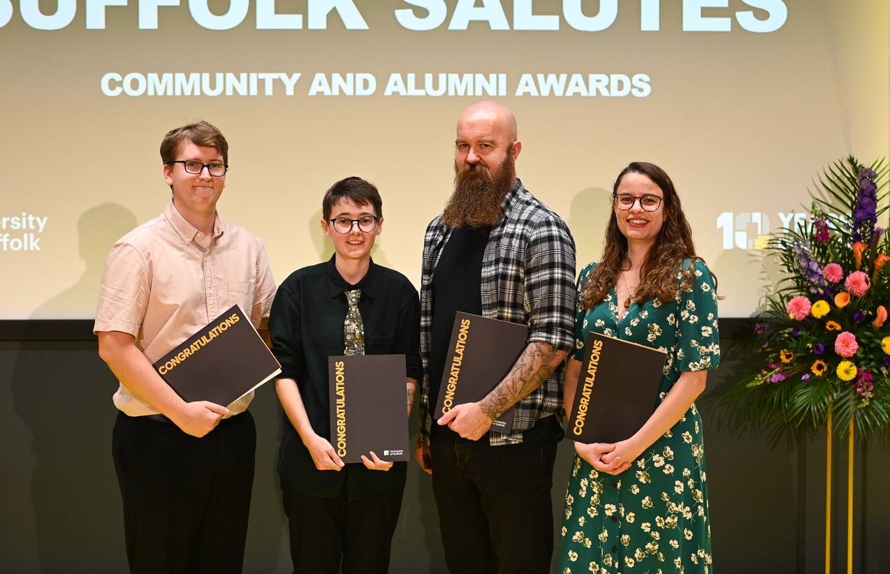 Four of the six Alumni Award winners standing on stage and smiling. They are holding their award certificates in a binder