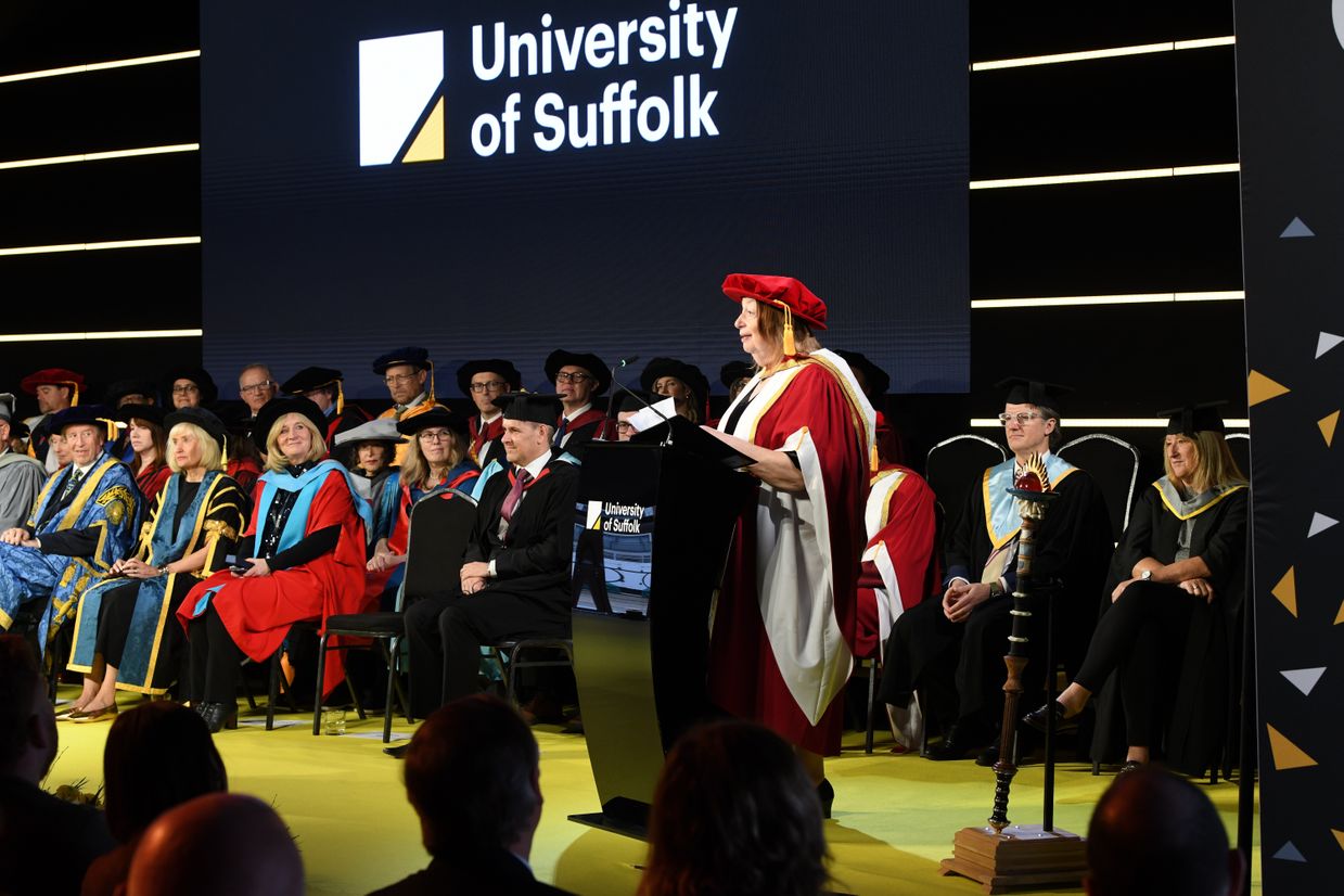 Alison Weir dressed in graduation robes and standing by a lectern on the stage at graduation. She is addressing students. Members of the congregation are seated behind