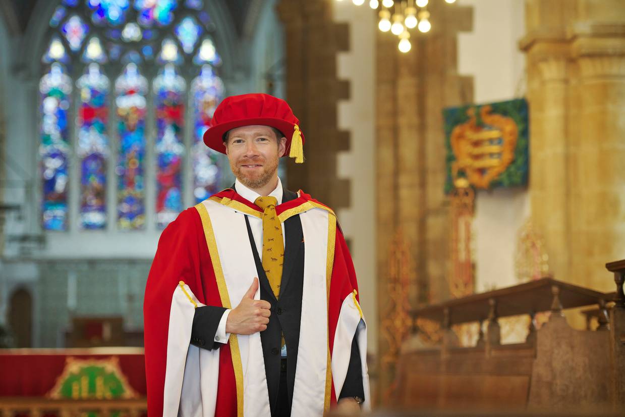 Zeb Soanes dressed in graduation robes making a thumbs up gesture and smiling