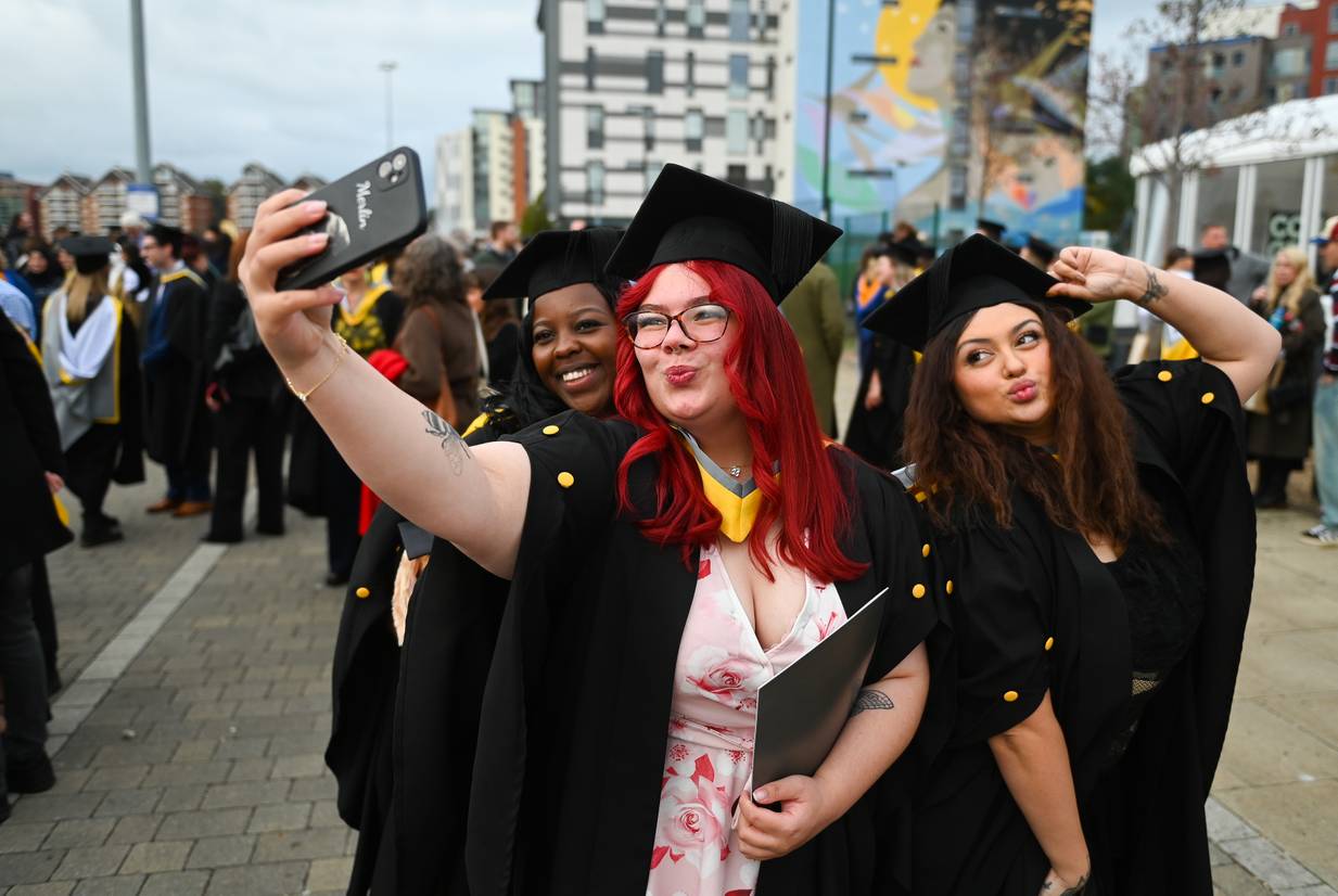 Three students on Ipswich waterfront dressed in graduation robes posing for a selfie