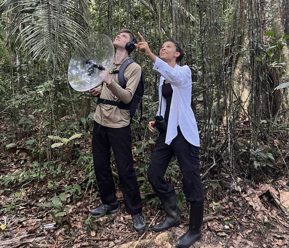 Students Dan Beckett and Leyla Huo standing in the Amazon Rainforest and looking up to the sky. Dan is using a directional microphone while Leyla is pointing into the sky at an animal