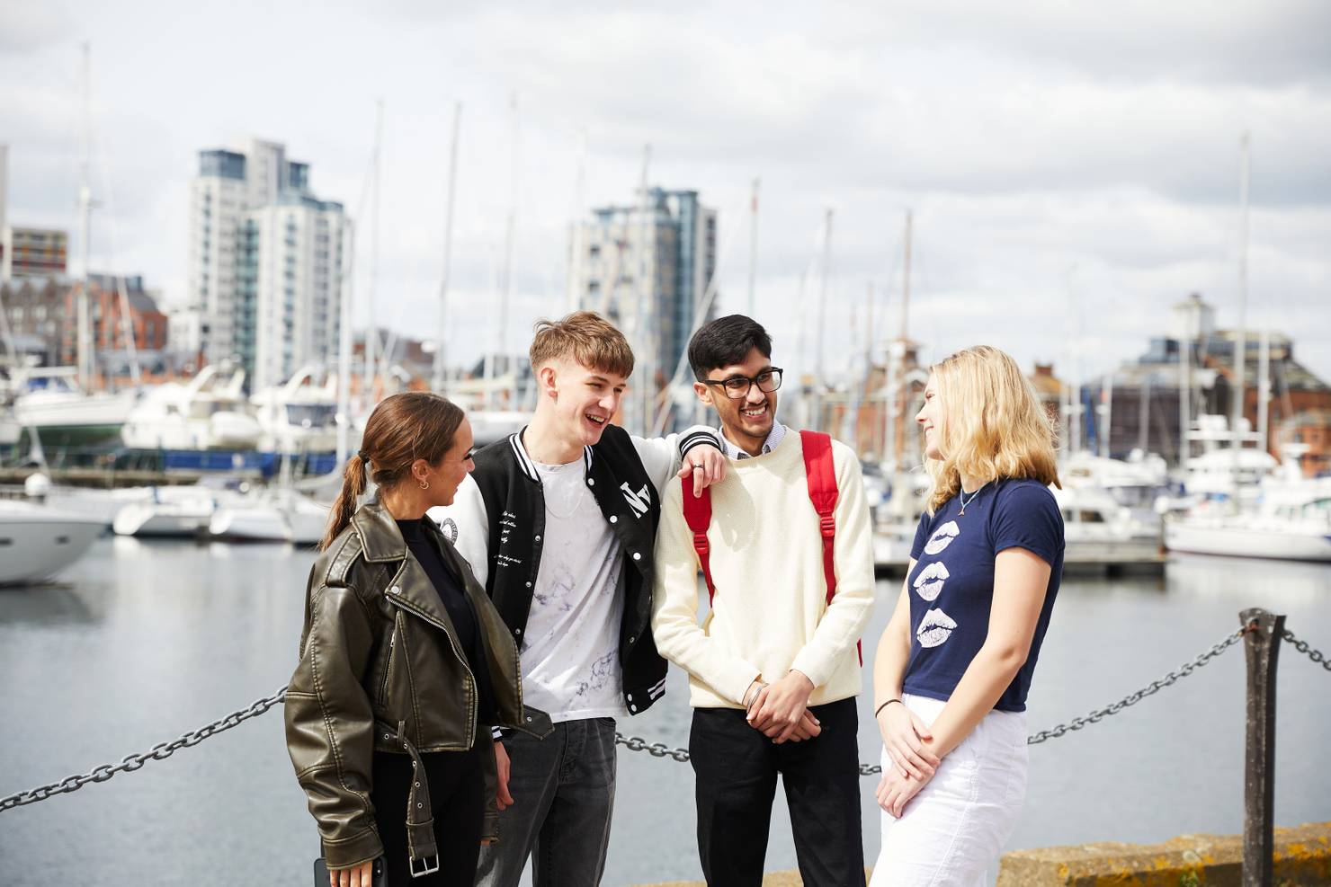 A group of four students standing on Ipswich Waterfront chatting and laughing. The marina is visible behind them