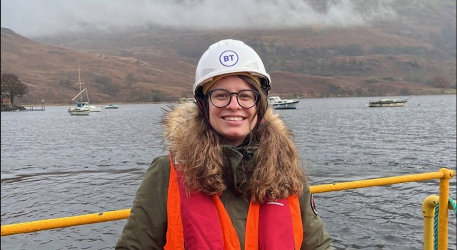 Digital apprenticeship graduate Xenia Zimmerman on site for BT in Scotland. Xenia is on a ship and smiling, wearing a hard hat and life jacket. The water of a Scottish loch is visible behind her