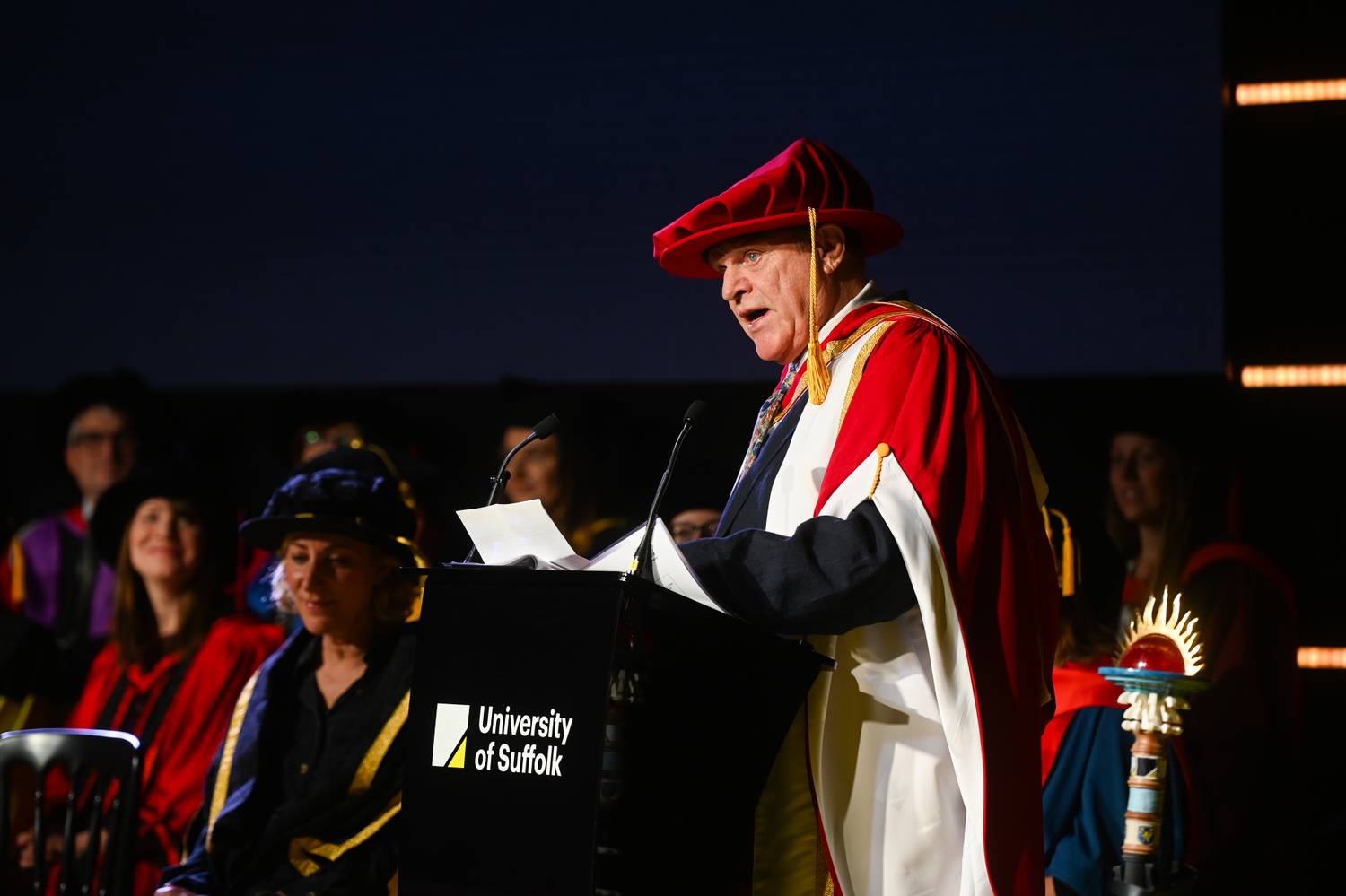 A photo of Charlie Haylock dressed in graduation robes, standing at the lectern delivering his speech