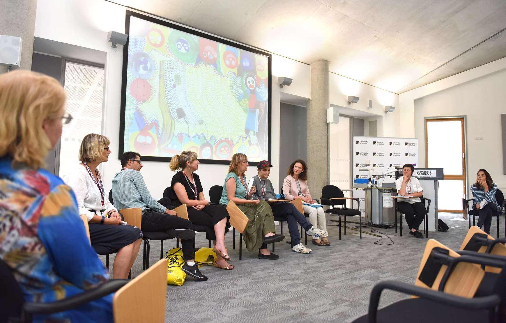 Delegates for the Storytelling Conference in one of the seminar room, seated at chairs arranged in a circle as part of a discussion. A piece of art is displayed on a projector behind.