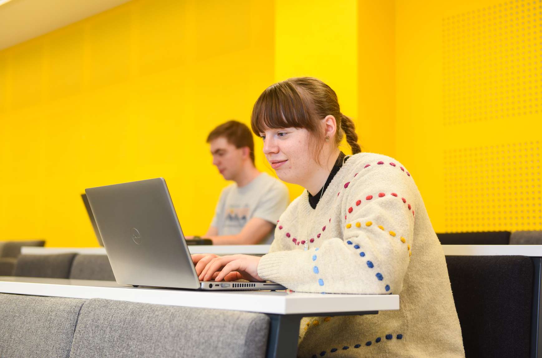 A student sitting in the lecture theatre typing at a laptop. Another student is visible in the row behind