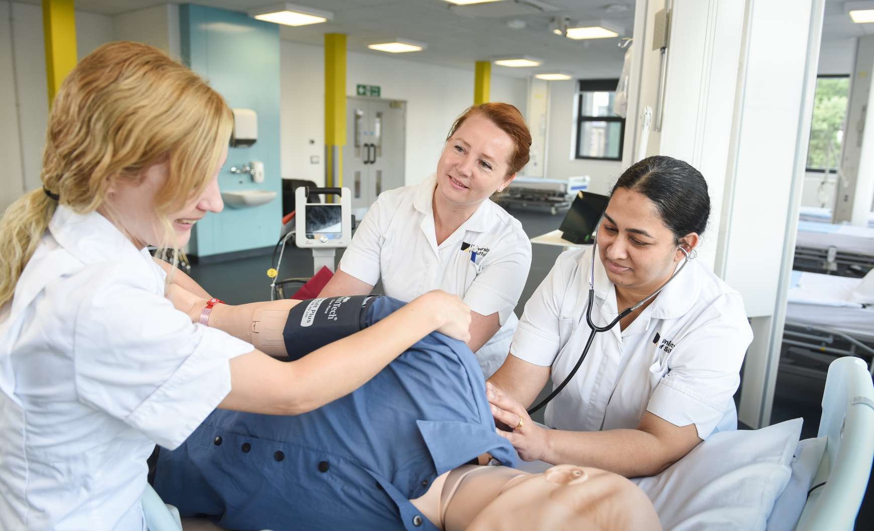 Three Adult Nursing students gathered around a hospital bed and working on some treatments on a training mannequin