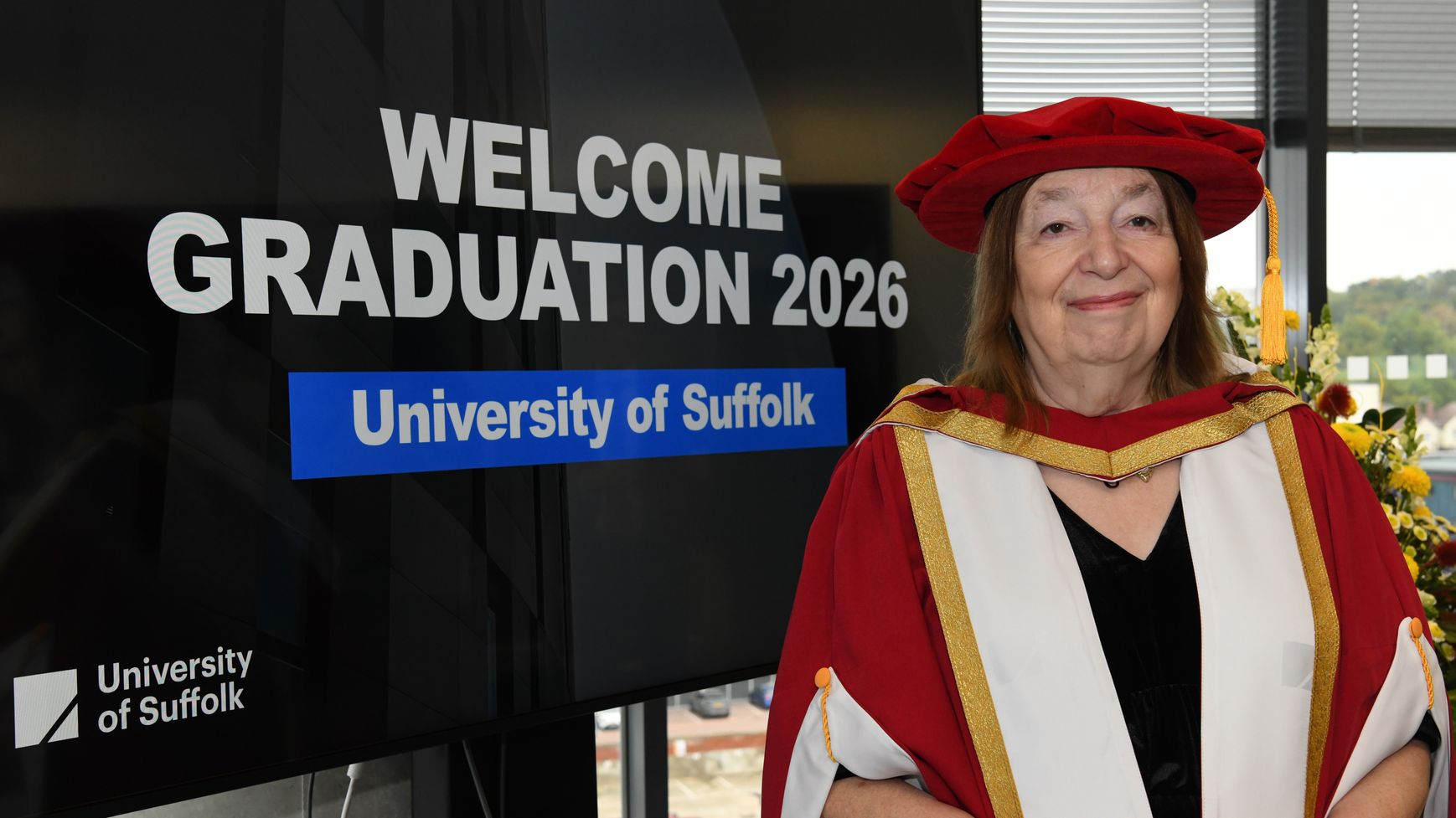 Dr Alison Weir dressed in graduation robes. She is standing next to a screen which says 'welcome to graduation 2026 - University of Suffolk'