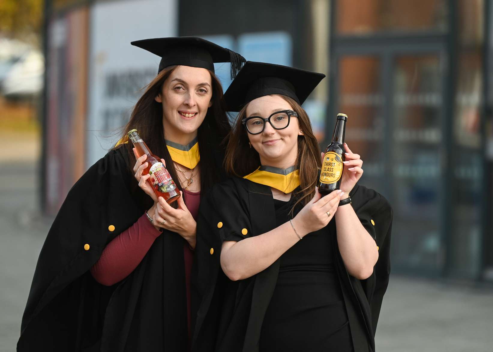 Business Management graduates Adele Harris (left) and Rebecca Mines dressed in their graduation robes and holding the brews they designed and made by Humber Doucy Brewing Company
