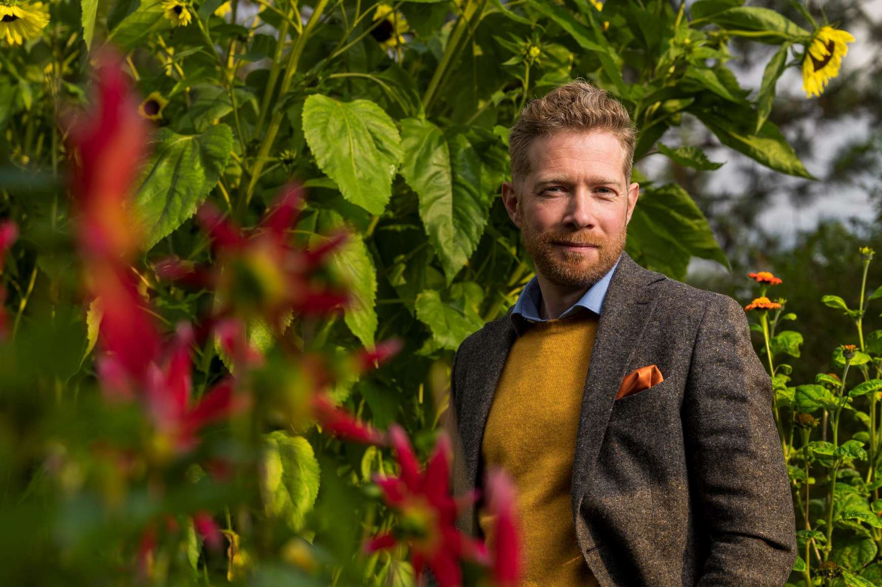 A head and shoulders image of Zeb Soanes standing by some shrubs