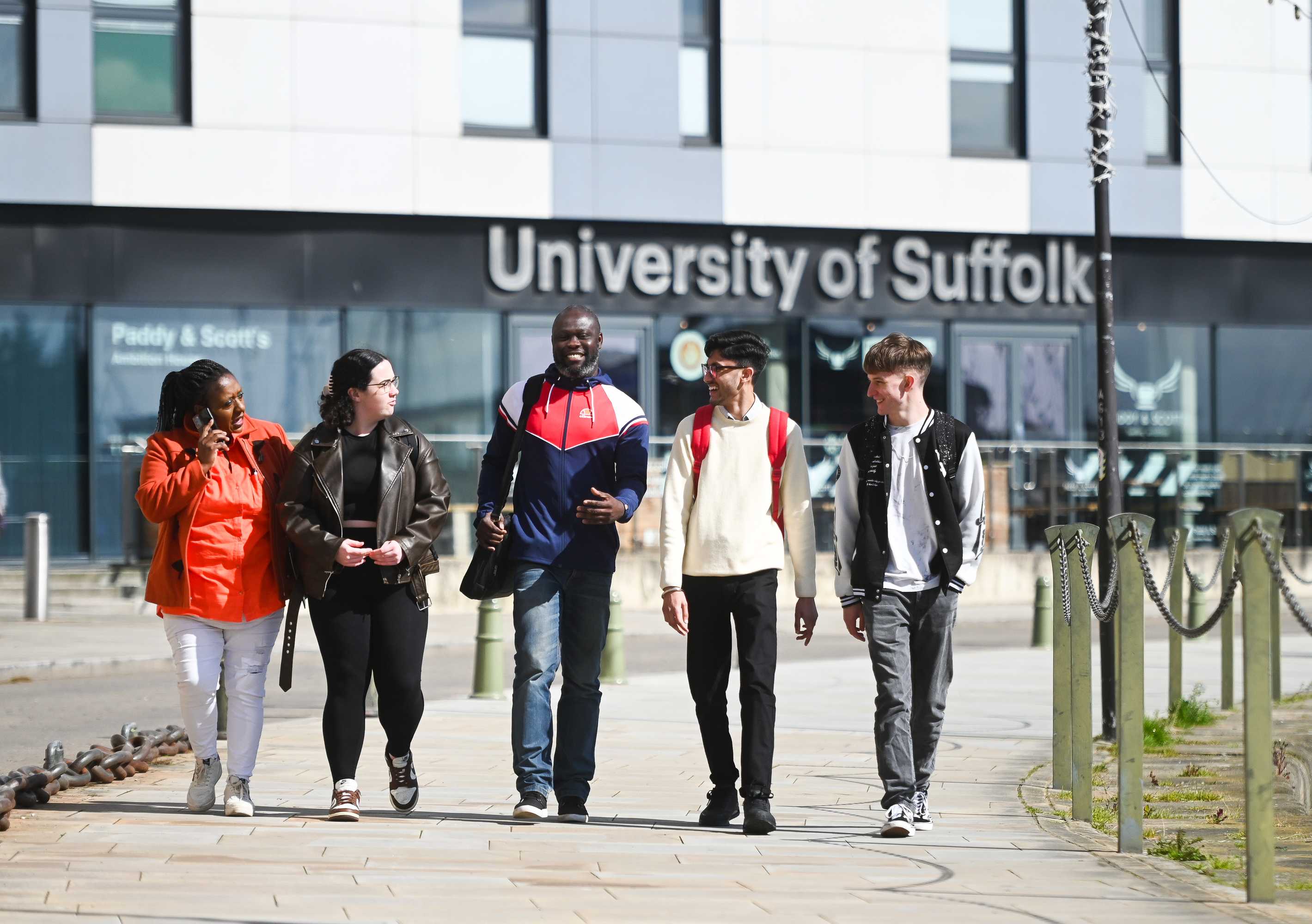 Group of five students chatting and walking in front of the Waterfront Building, with the University of Suffolk building sign behind them.