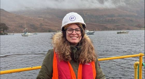 Digital apprenticeship graduate Xenia Zimmerman on site for BT in Scotland. Xenia is on a ship and smiling, wearing a hard hat and life jacket. The water of a Scottish loch is visible behind her