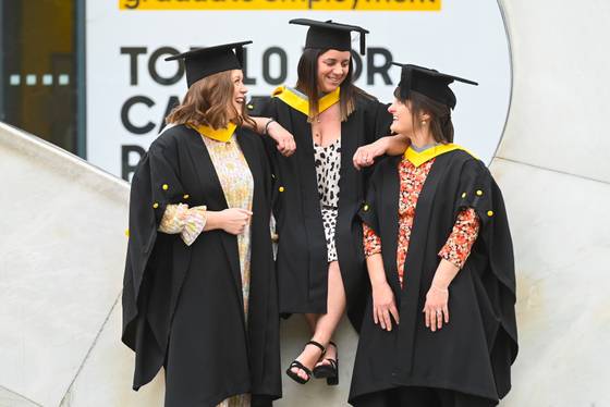 Three students in gradation robes from 2023. One is sitting on the question mark statue while the other two are talking to her.