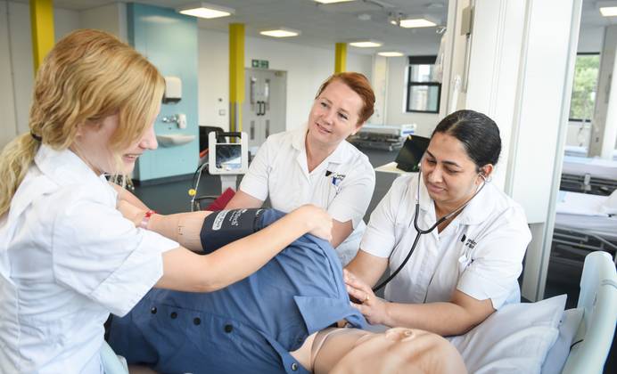 Three Adult Nursing students gathered around a hospital bed and working on some treatments on a training mannequin