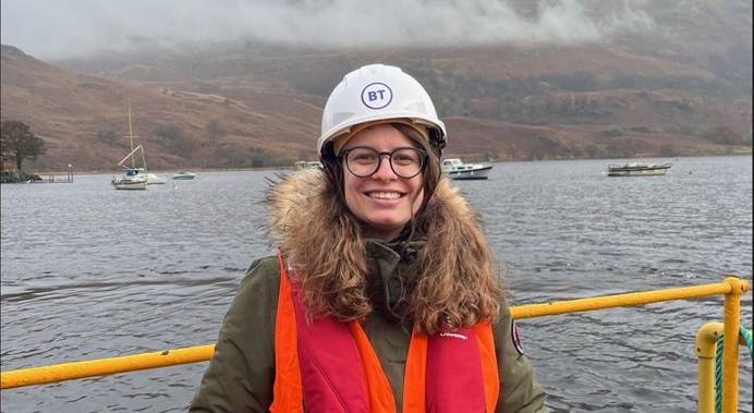 Digital apprenticeship graduate Xenia Zimmerman on site for BT in Scotland. Xenia is on a ship and smiling, wearing a hard hat and life jacket. The water of a Scottish loch is visible behind her