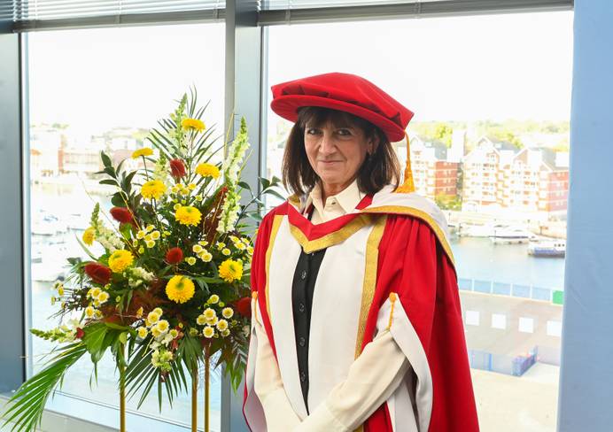 Emma Freud dressed in graduation robes and standing beside some flowers. Ipswich marina is visible out of the window behind her
