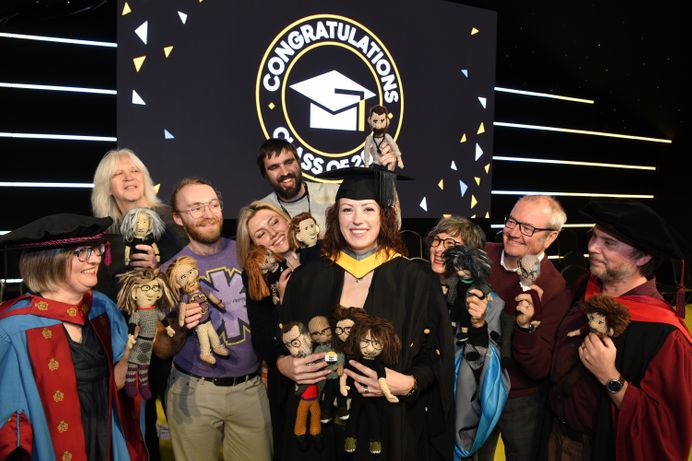 Gemma Upson dressed in graduation robes and holding some of the caricatures she made. She is surrounded by arts technicians and tutors who are holding the caricatures of themselves Gemma made