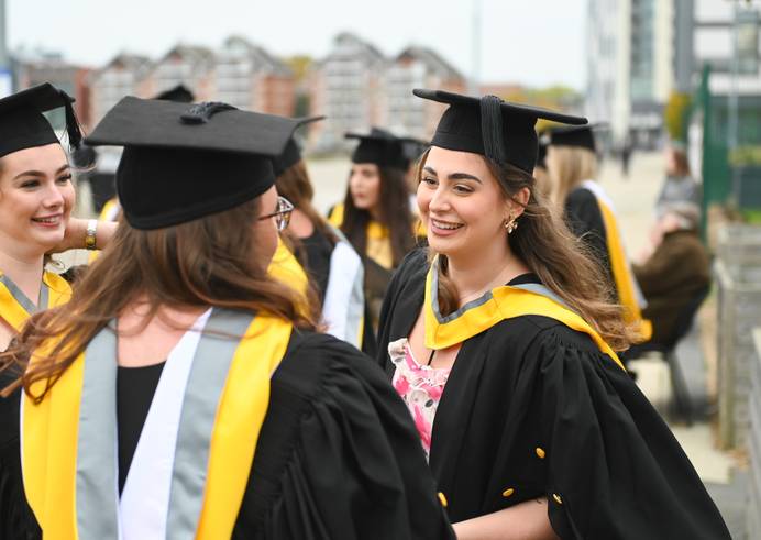 Three students dressed in graduation robes, talking and smiling