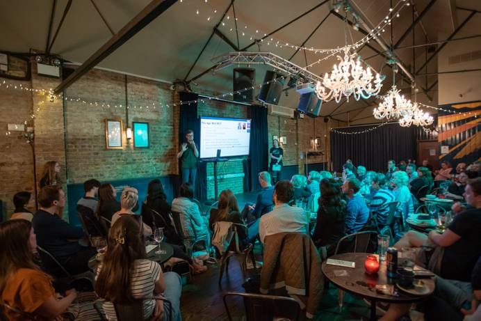A pint of Science event elsewhere in the UK. A researcher is standing on a small stage with a screen visible behind and presenting to a pub room full of listening participants