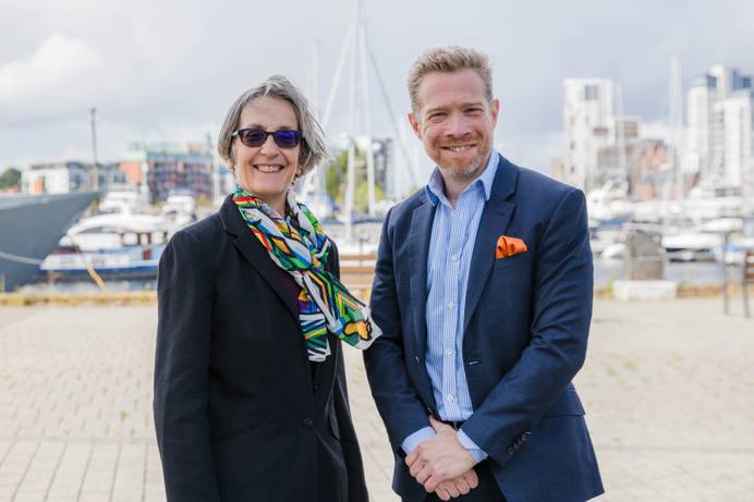 The outgoing Chancellor Helen Pankhurst (left) and new Chancellor Zeb Soanes (right) standing on Ipswich waterfront and smiling