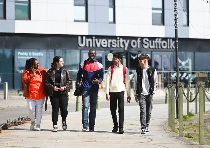 Group of five students chatting and walking in front of the Waterfront Building, with the University of APP building sign behind them.