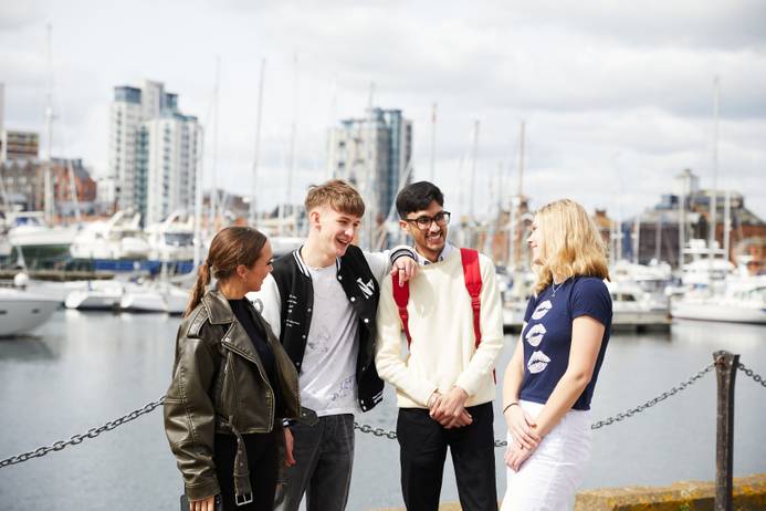 A group of four students standing on Ipswich Waterfront chatting and laughing. The marina is visible behind them