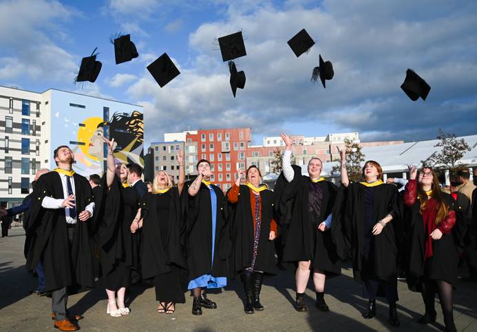 Students at the 2024 graduation ceremonies standing outside on the waterfront and tossing their mortar board hats into the air in celebration