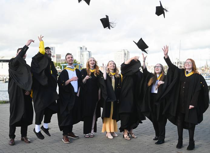 Students at the 2025 graduation ceremonies dressed in graduation robes. They sre standing on the waterfront in a line and tossing their caps in the air in celebration