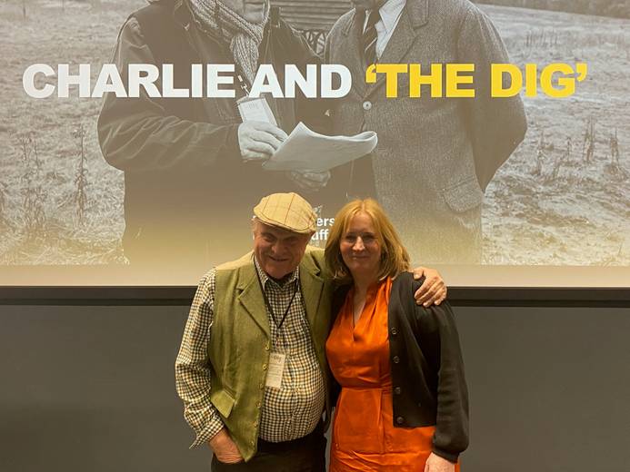 Charlie Haylock (left) and Dr Amanda Hodgkinson (right) standing side by side and smiling at the open lecture event. The words 'Charlie and The Dig' are visible on the screen behind them