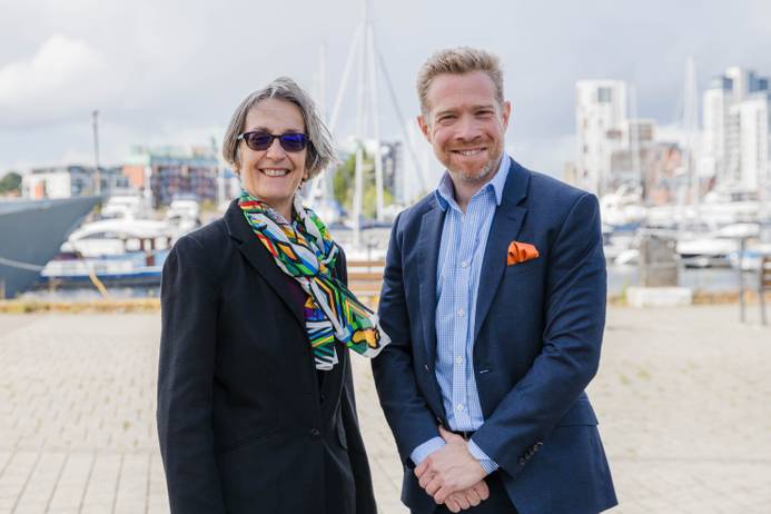 The outgoing Chancellor Helen Pankhurst (left) and new Chancellor Zeb Soanes (right) standing on Ipswich waterfront and smiling