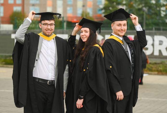 Three students wearing graduation caps and gowns tipping their caps