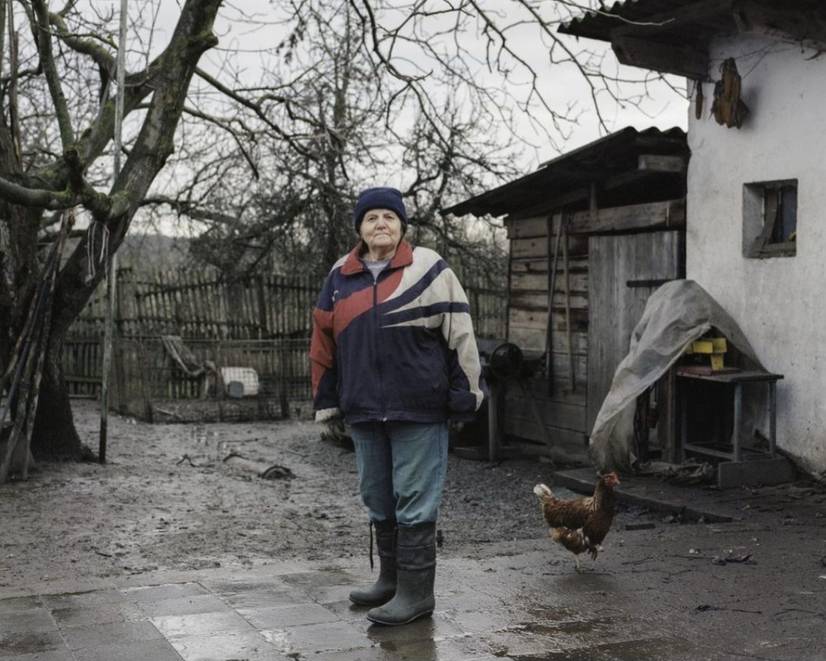 A photograph of a woman in coat and wellies standing in her back yard. The ground is damp and muddy and a chicken roams the ground