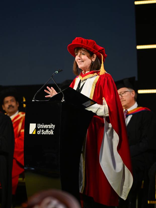Emma Freud dressed in graduation robes and standing at a lectern, addressing students.