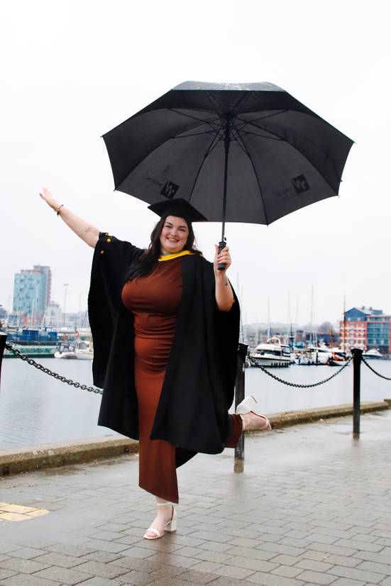 SU President Zoe Saunders dressed in graduation robes and standing under umbrella on Ipswich waterfront