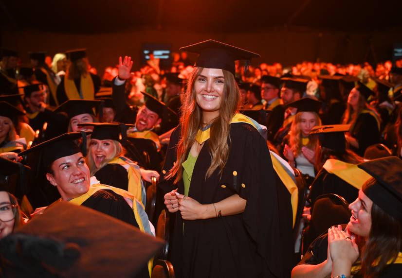 A student dressed in graduation robes inside the marquee, surrounded by course mates, smiling
