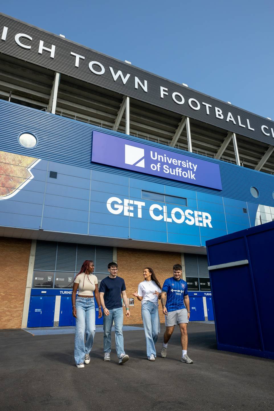 Four students with Sir Alf Ramsey stand dominating the background. University of Suffolk logo and sign 